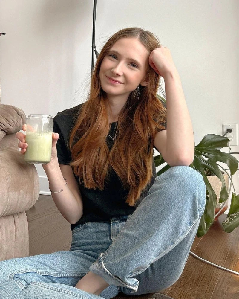 A person with long reddish-brown hair sits on the floor, wearing a black t-shirt and blue jeans. They hold a glass of green smoothie, smiling softly. A plant is visible in the background.